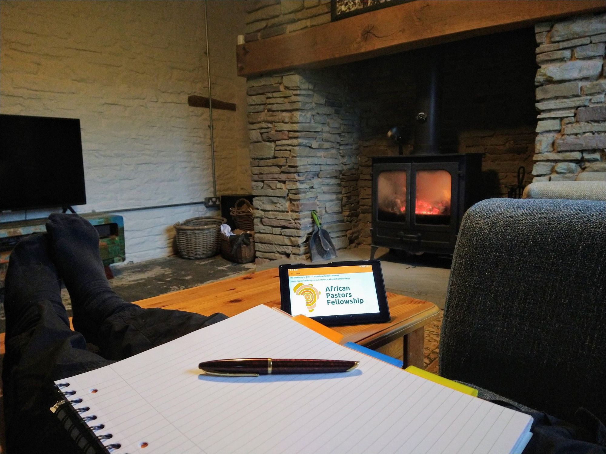 A lit log burning stove in a fire place, with a table in front of it.  In the foreground is a notepad and pen resting on a person's outstretched legs.