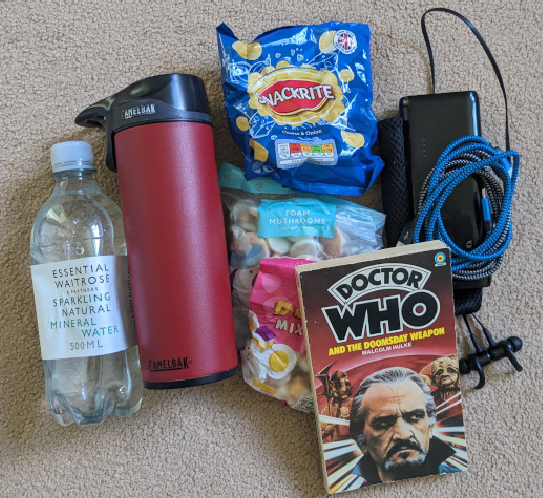 Pile with a bottle of water, flask of tea, book, crisps, bags of sweets, backup battery and cables and bluetooth headset.