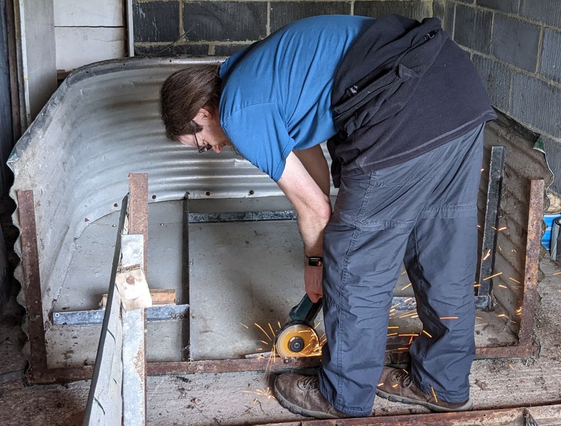 Photograph of a man leaning over and using an angle grinder on metalwork that's resting on the floor. There are orange sparks flying around the man's legs.
