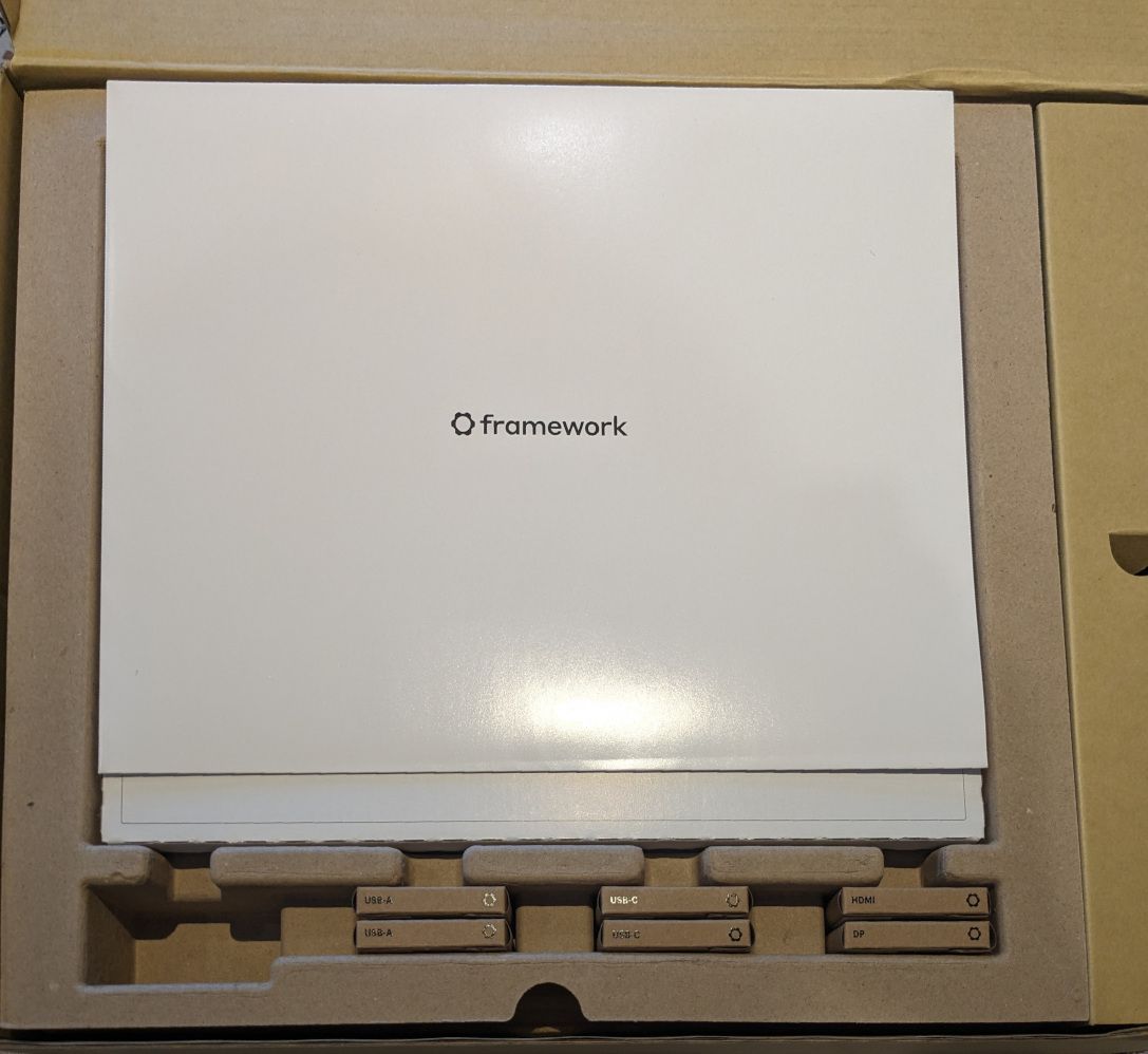 Photograph of boxed Framework 16 laptop.  Small brown boxes containing the expansion cards are shown at the bottom.  At the top is a white cardboard envelope.  The laptop is not yet visible.