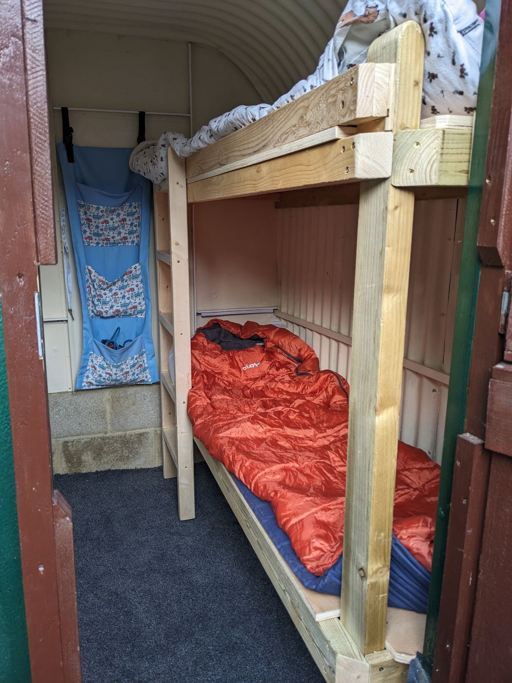 Photograph showing a blue carpeted floor. On the right hand side are two bunk beds made out of wood, with a ladder at the far end. The bottom bunk has an orange sleeping bag on it, with blankets on the top bunk.