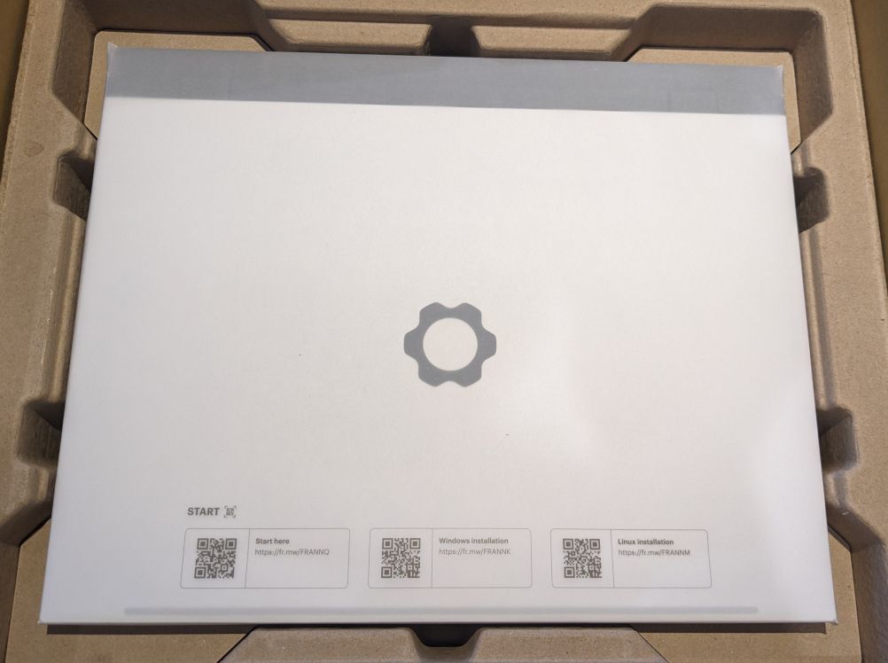 The laptop wrapped in tracing paper, placed in the cardboard surround.  Framework's logo looks pale due to the paper, and the laptop itself isn't shiny for the same reason.