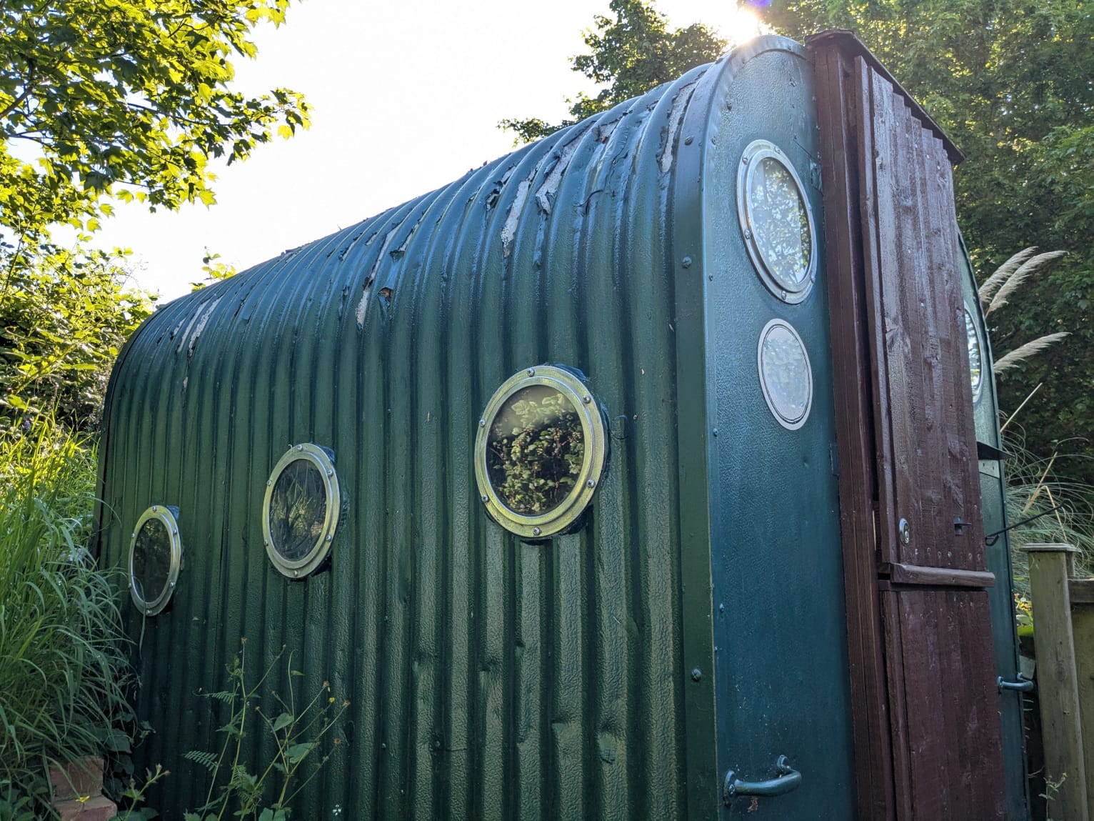 Corrugated metal hut painted a dark green. The paint is peeling on the top. There's a brown wooden door on the front, and circular glass portholes on the side wall and either side of the door. Also used for the banner image at the top of this post.
