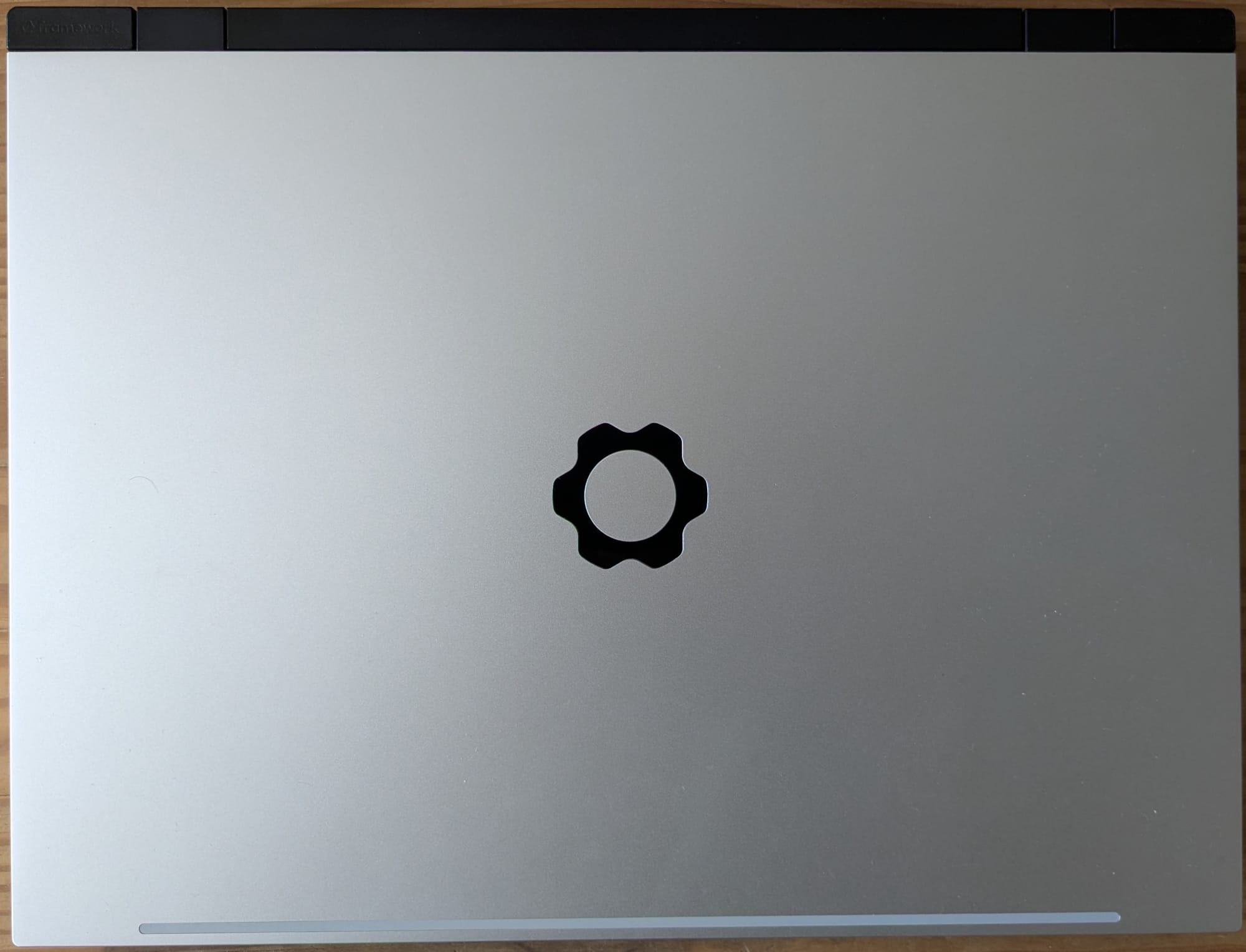 Photograph showing the Framework 16 laptop with the expansion bay shell installed. The laptop is not as deep as it is with the dedicated GPU installed. The laptop is primarily a silver metal with a shiny cog symbol (the Framework logo) in the centre.