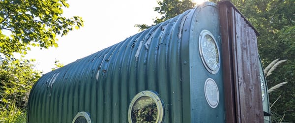 Corrugated green metal hut, with a brown wooden door and glass portholes.