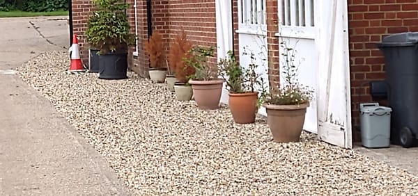 Photograph showing the side of a brick building, with a concrete path on the left and large pebbles by the building on the right.  There are white framed windows with white slatting beneath.
